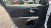 Close-up of a car door interior featuring a black textured panel, chrome door handle, and window controls; outside is a parking lot with trucks and cars visible.
