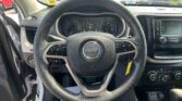 Driver's-eye view of a Jeep interior with steering wheel and instrument cluster, center console, and parked cars seen through the windshield