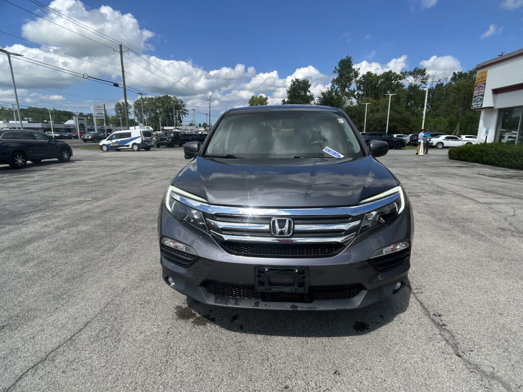 Close-up, front view of a gray Honda SUV in a parking lot with other cars and a dealership in the background on a sunny day with blue sky and clouds.