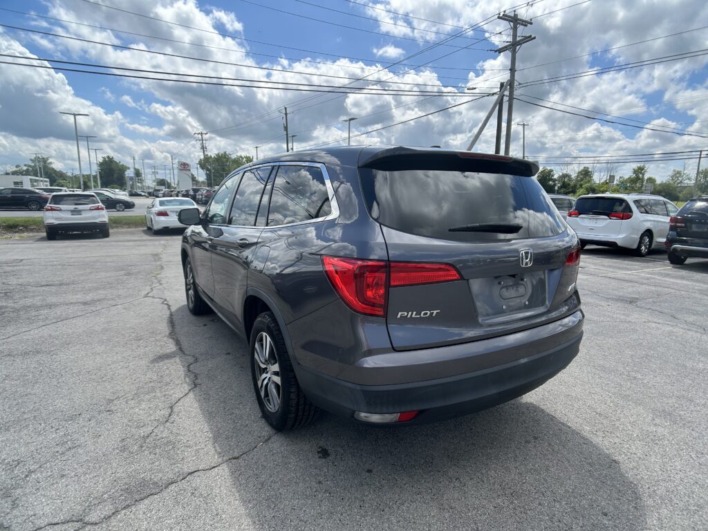 Rear view of a gray Honda Pilot SUV parked in a car dealership lot with other vehicles and power lines overhead against a partly cloudy sky.