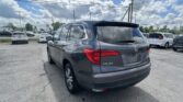 Rear view of a gray Honda Pilot SUV parked in a car dealership lot with other vehicles and power lines overhead against a partly cloudy sky.