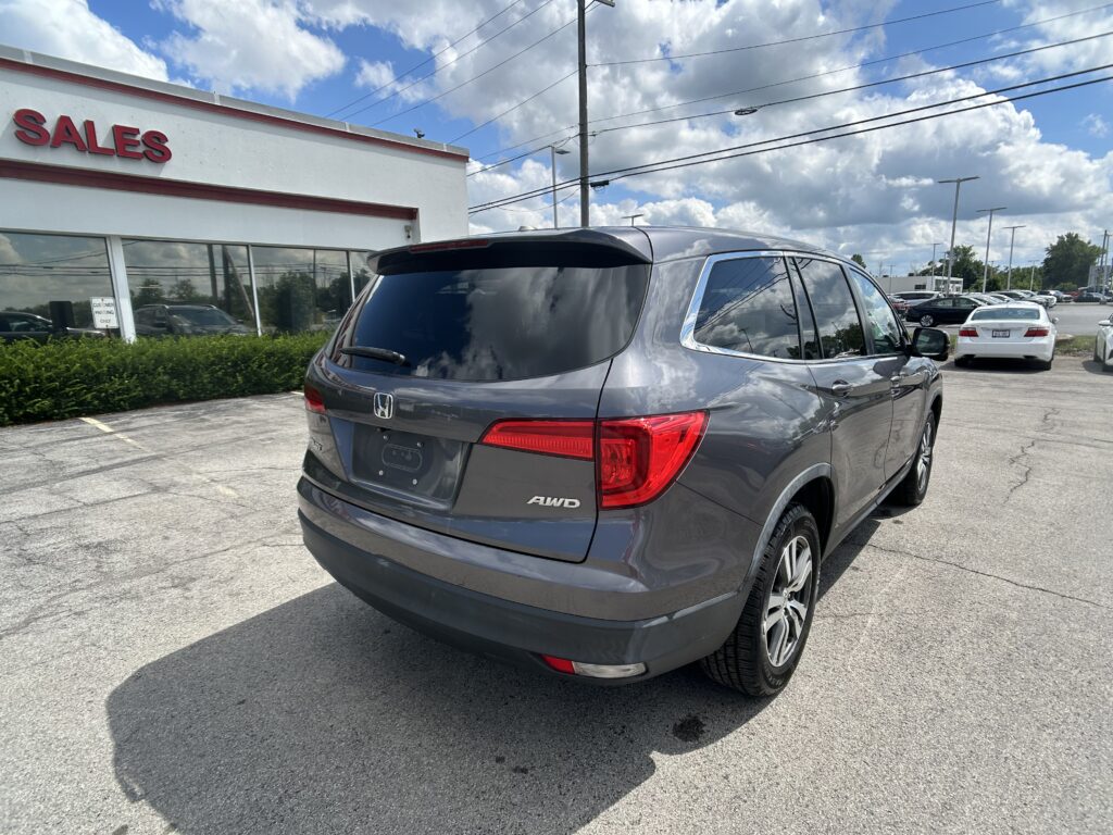 Rear three-quarter view of a gray Honda SUV in a dealership parking lot in front of a white building with a red 'SALES' sign, blue sky above.