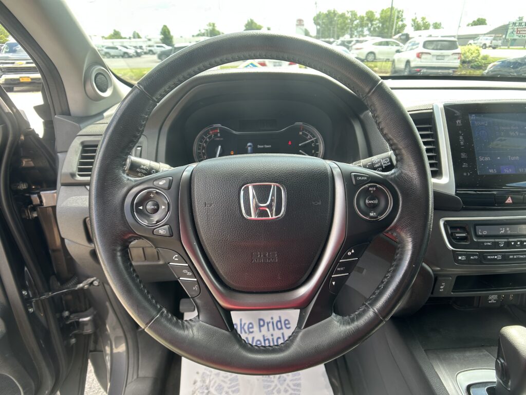Driver's view of a black leather steering wheel with Honda emblem, instrument cluster, and center console, looking out at a busy parking lot through the windshield.