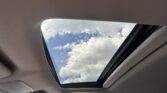 Inside a car, the panoramic sunroof is open, showing a blue sky with white clouds and raindrops on the glass.