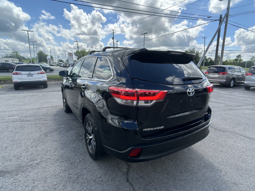 Rear three-quarter view of a black Toyota Highlander parked in a dealership lot with other cars and power lines overhead under a cloudy blue sky.”, “Black Toyota Highlander in a parking lot, seen from the rear side with glossy finish and red taillights.”, “Row of cars at a dealership lot with a black Highlander in the foreground and a cloudy sky.”, “Close-up of the back of a black Highlander SUV, showing the Highlander badge and taillights.”, “Vehicle lot scene with multiple cars and tall power poles under a partly cloudy sky, foreground vehicle is a black SUV.”
