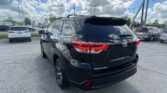 Rear three-quarter view of a black Toyota Highlander parked in a dealership lot with other cars and power lines overhead under a cloudy blue sky.”, “Black Toyota Highlander in a parking lot, seen from the rear side with glossy finish and red taillights.”, “Row of cars at a dealership lot with a black Highlander in the foreground and a cloudy sky.”, “Close-up of the back of a black Highlander SUV, showing the Highlander badge and taillights.”, “Vehicle lot scene with multiple cars and tall power poles under a partly cloudy sky, foreground vehicle is a black SUV.”