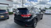 Rear view of a black SUV at a car dealership lot, with a white building, other vehicles, and a cloudy blue sky.