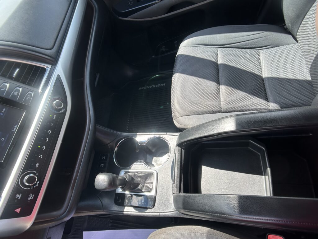 Interior of a car showing the center console with climate controls, gear shifter, and cup holders, next to a gray patterned front passenger seat.
