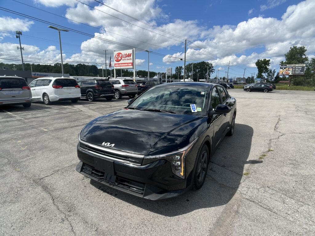 Black Kia SUV in a busy car lot with several vans and SUVs, an EZ Loan Auto sign, and a blue sky with clouds overhead.