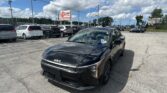 Black Kia SUV in a busy car lot with several vans and SUVs, an EZ Loan Auto sign, and a blue sky with clouds overhead.