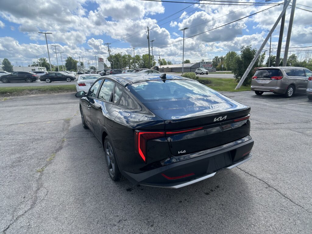 Rear view of a black Kia sedan in a crowded parking lot, with power lines and a blue sky with white clouds nearby.