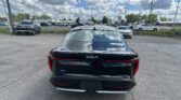 Rear of a black Kia sedan in a dealership parking lot, with blue sky and clouds reflecting off the trunk and other cars in the background.] | Note: Keep under 125 chars for actual ALT text in code- here are options.
