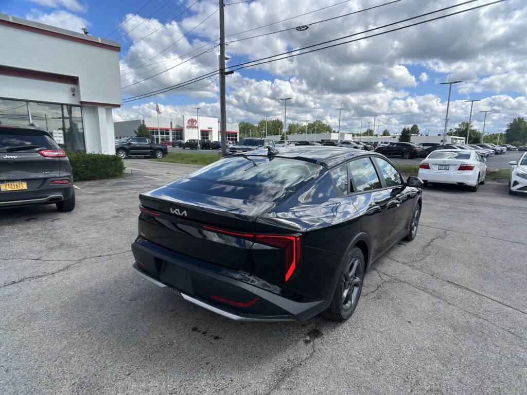 Rear three-quarter view of a black Kia SUV in a car dealership lot, with a Toyota showroom in the background and a cloudy blue sky above.