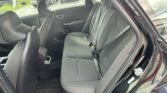 Interior of a car's rear passenger row with dark fabric seats, three seat belts, headrests, and a door panel; view through an open rear door to parked cars outside.