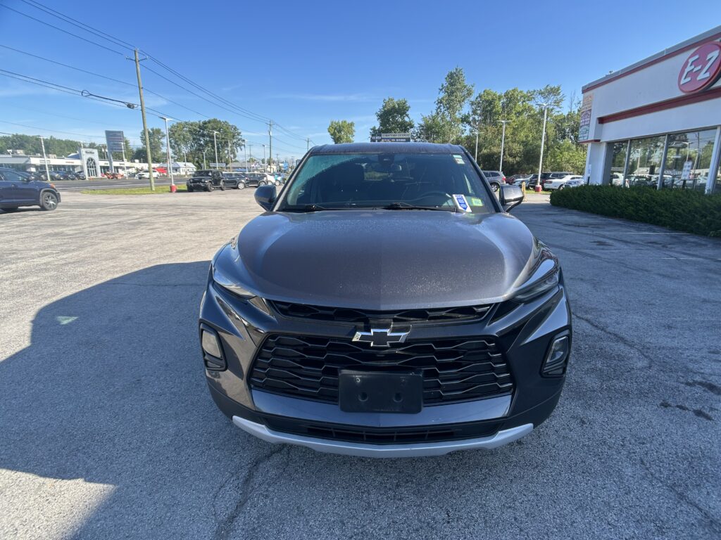 Close-up of the front of a dark gray Chevrolet SUV parked in a car dealership lot, with a building and trees in the background.