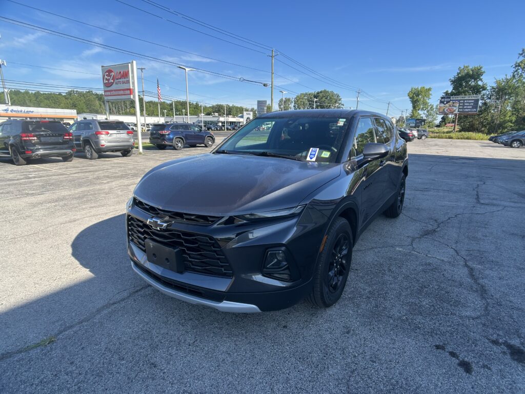 Front-right view of a dark gray Chevrolet SUV in a busy car dealership lot on a sunny day, with other vehicles and signs in the background.