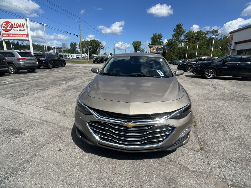 Front view of a gray Chevrolet sedan in a busy used-car lot with an E-Z Loan Auto Sales sign on the left and other vehicles parked nearby under a bright blue sky.