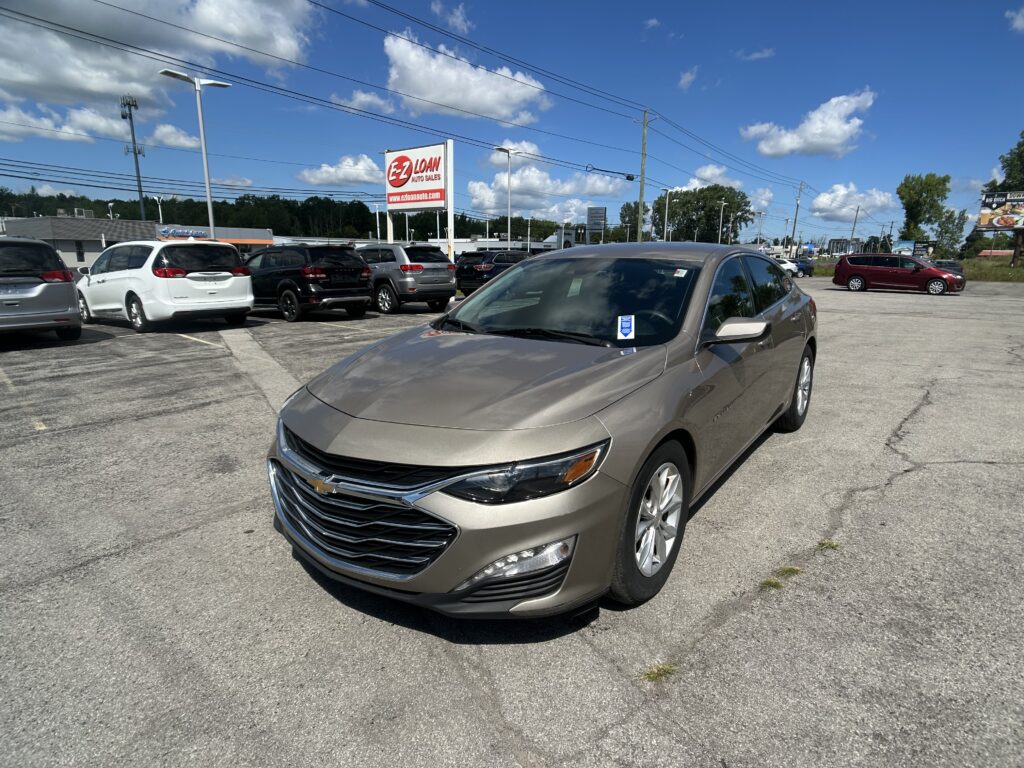 Front-right view of a tan Chevrolet sedan in a busy car dealership lot with other SUVs and a red EZ LOAN Auto Sales sign against a blue sky.
