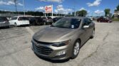 Front-right view of a tan Chevrolet sedan in a busy car dealership lot with other SUVs and a red EZ LOAN Auto Sales sign against a blue sky.