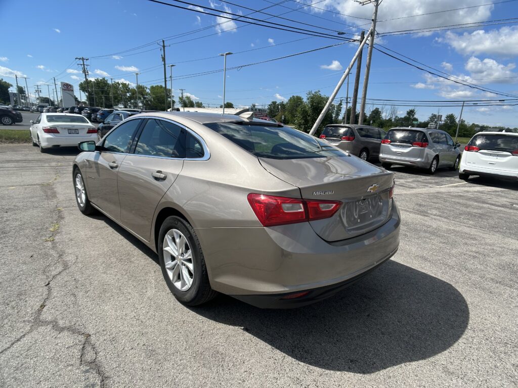 Tan Chevrolet Malibu in the foreground at a car dealership, with a row of cars behind and a blue sky above.