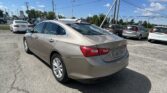 Tan Chevrolet Malibu in the foreground at a car dealership, with a row of cars behind and a blue sky above.