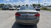 Rear view of a gray Chevrolet sedan in a dealership parking lot with other cars and a blue sky filled with clouds above.