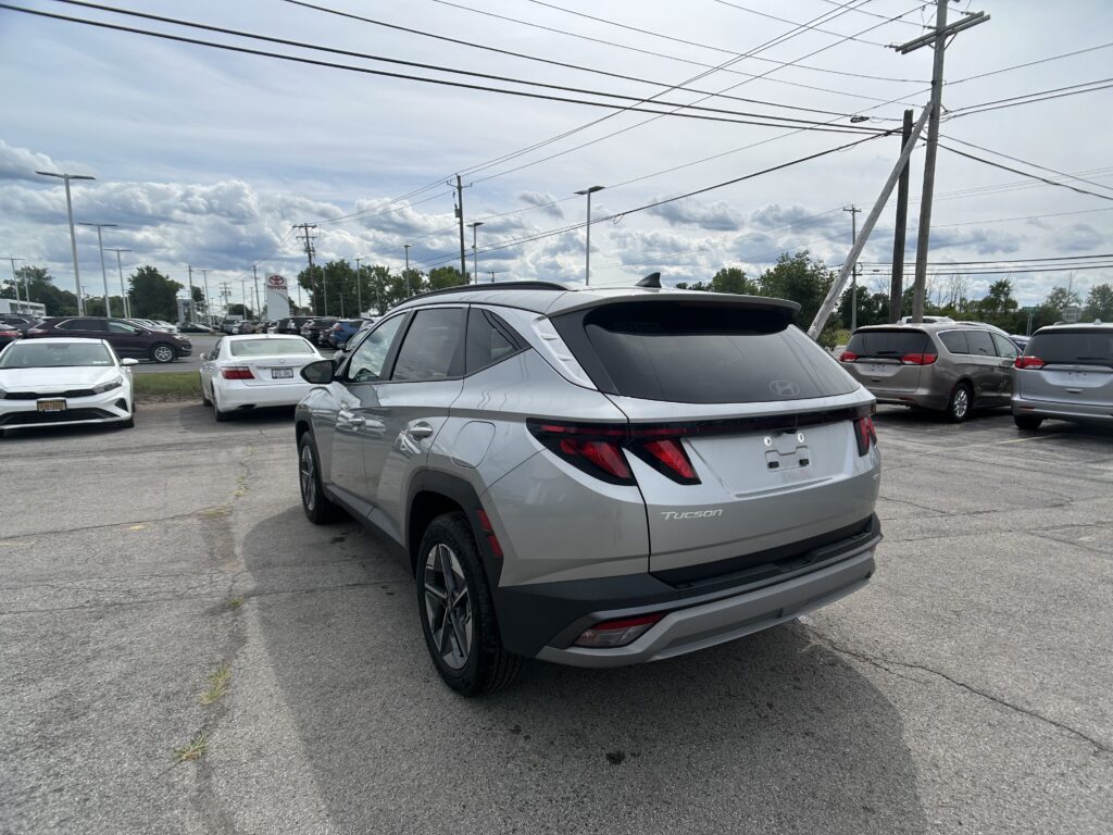 Silver Hyundai Tucson in the foreground of a crowded car dealership lot with many parked vehicles and power lines overhead in a cloudy sky.