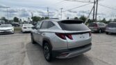 Silver Hyundai Tucson in the foreground of a crowded car dealership lot with many parked vehicles and power lines overhead in a cloudy sky.