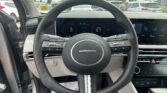 Car interior view from the driver seat showing a digital dashboard and steering wheel, with a dealership lot visible through the windshield.