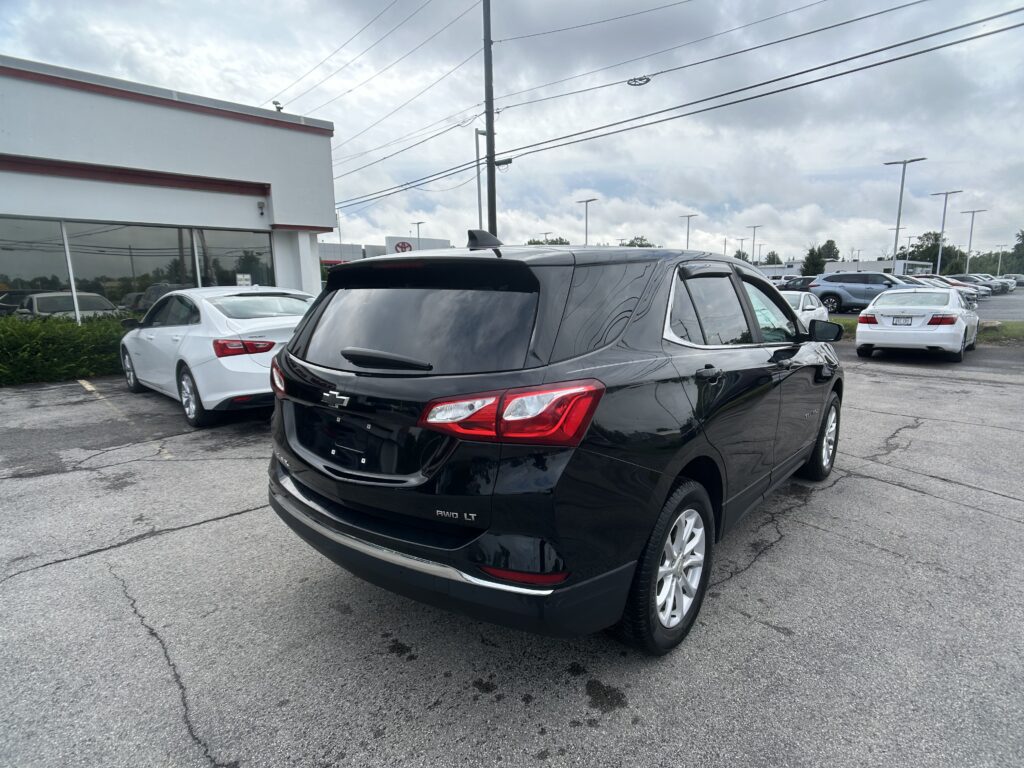 Black SUV in the foreground at a car dealership lot, with white cars and a white showroom building on the left under a cloudy sky.