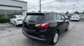 Black SUV in the foreground at a car dealership lot, with white cars and a white showroom building on the left under a cloudy sky.