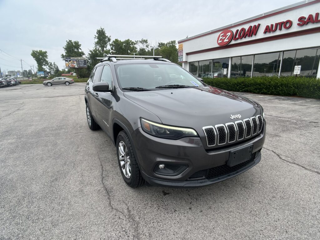 Front-left angle of a gray Jeep SUV with roof rails parked in a lot, in front of an E-Z Loan Auto dealership building.