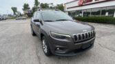 Front-left angle of a gray Jeep SUV with roof rails parked in a lot, in front of an E-Z Loan Auto dealership building.