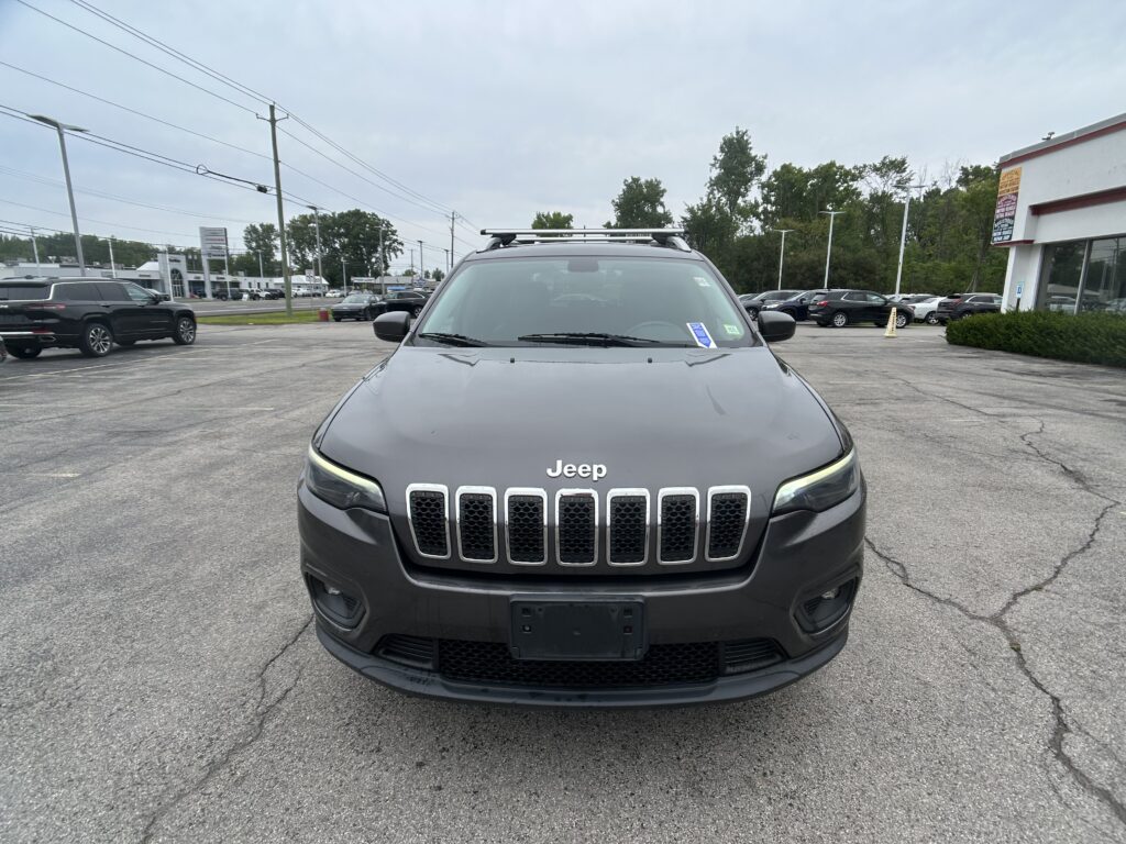Front view of a gray Jeep SUV with a roof rack in a dealership parking lot at a wide-angle shot.