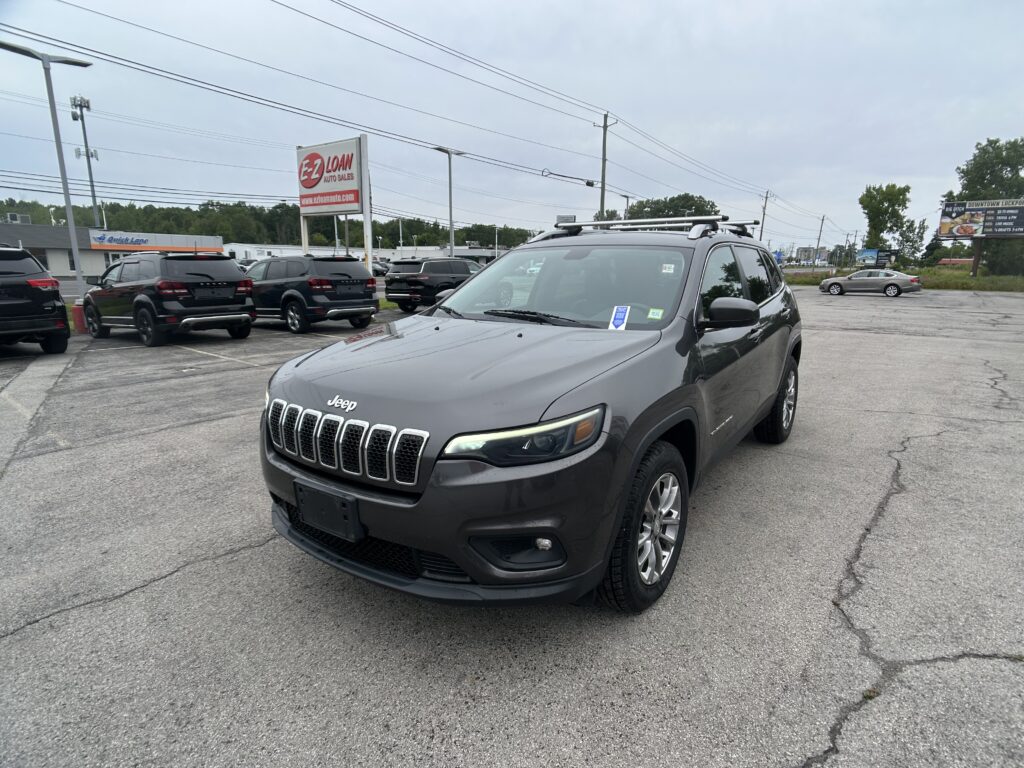 Gray Jeep SUV in the foreground at a car dealership lot, with other vehicles and signs in the background.