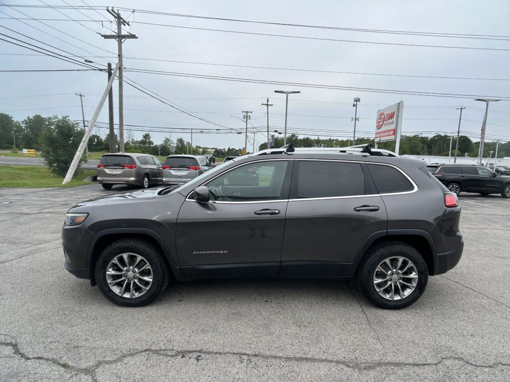 Gray Jeep Grand Cherokee in a parking lot with other vehicles and a power line-filled sky in the background, dealership sign visible in distance