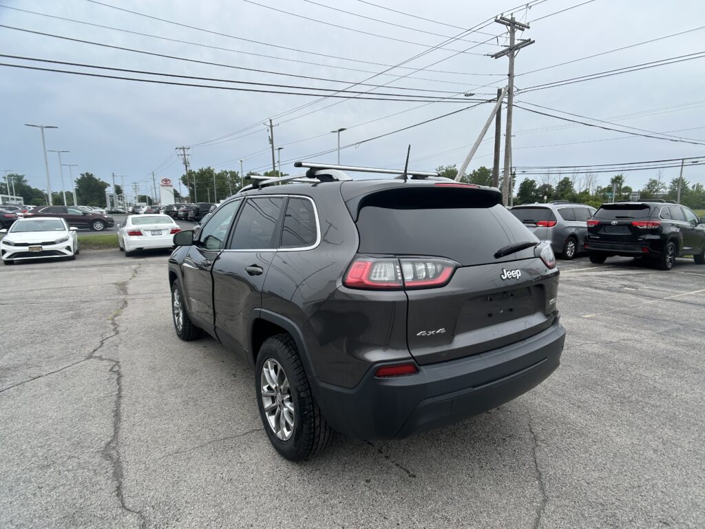 Rear three-quarter view of a gray Jeep with roof rack in a busy parking lot, rows of cars and overhead power lines in the background.