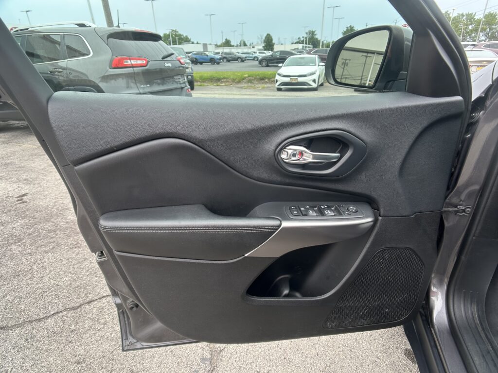 Interior car door panel with window controls, viewed from the front passenger seat. Outside: a parking lot with cars and a white sedan visible in the mirror.