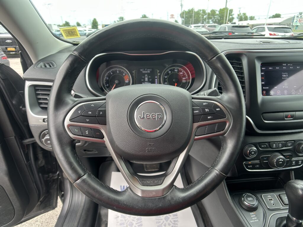 Driver's view from the seated position inside a Jeep, steering wheel centered with instrument cluster visible behind and a touchscreen infotainment system to the right; parking lot seen through the windshield.