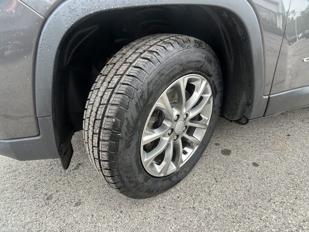 Close-up of a wet car tire and silver alloy wheel on a gray vehicle, showing rain droplets on the fender and pavement nearby.