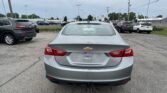 Rear view of a silver Chevrolet Malibu in a busy outdoor car dealership lot, with many parked cars and a Toyota sign in the distance.