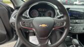 View from the driver’s seat showing a Chevrolet steering wheel, dashboard gauges, and a touchscreen, with a car lot outside the windshield.