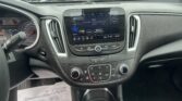 Interior of a car dashboard with a touchscreen infotainment system displaying FM radio and Aerosmith playing, parked in a lot seen through the windshield.