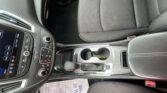 Interior view of a car's front cabin showing the center console with touchscreen display, climate controls, gear shifter, cup holders, and part of the passenger seat with a paper floor mat nearby.