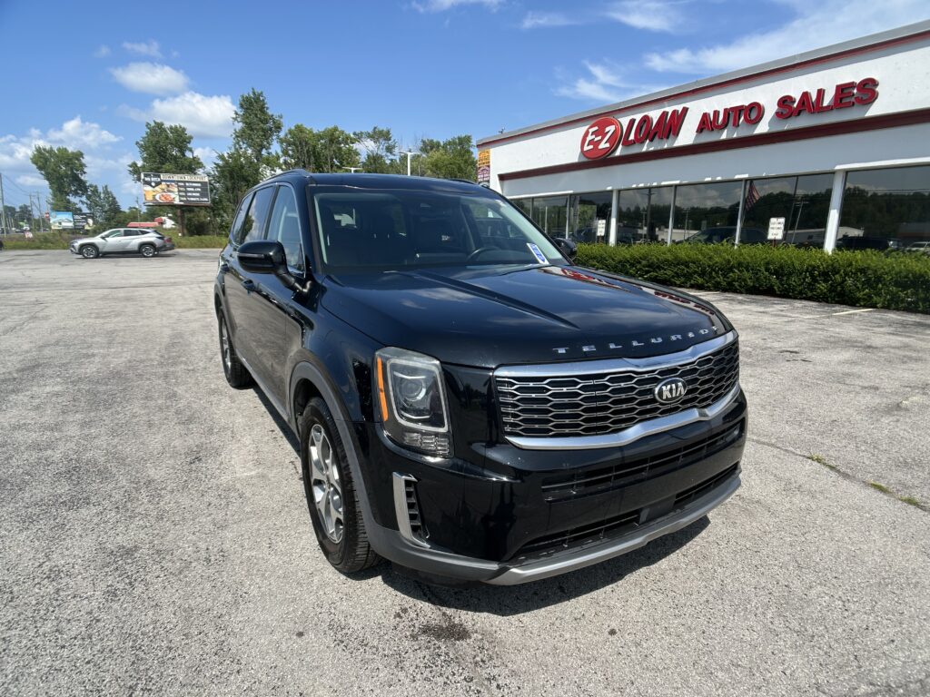 Black Kia Telluride in the foreground outside EZ Loan Auto Sales showroom on a sunny day with blue sky and other cars in the lot