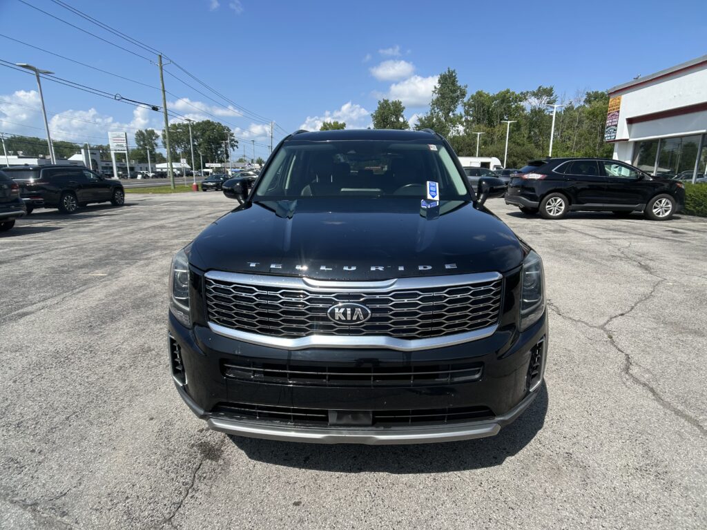 Front view of a black Kia Telluride parked in a dealership lot with other cars and a white building to the right, under a blue sky.