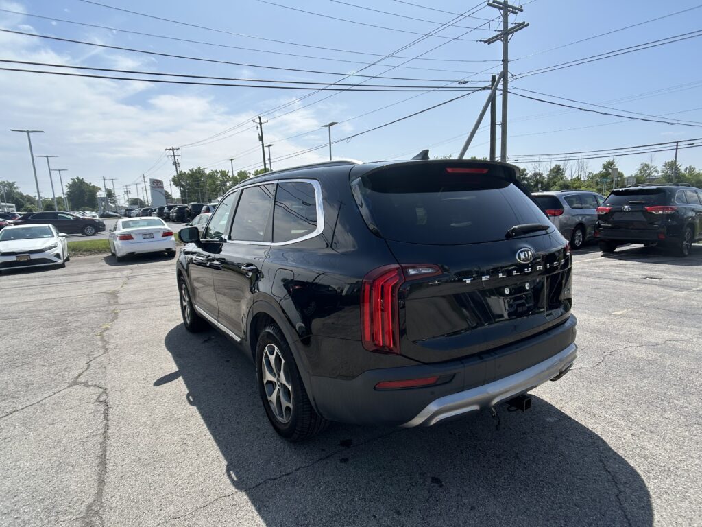 Rear three-quarter view of a black Kia Telluride SUV in a crowded car dealership lot, power lines overhead.