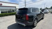 Black Kia Telluride SUV viewed from the rear‑three‑quarter, parked in a dealership lot next to a white building with red trim and other cars in the background.