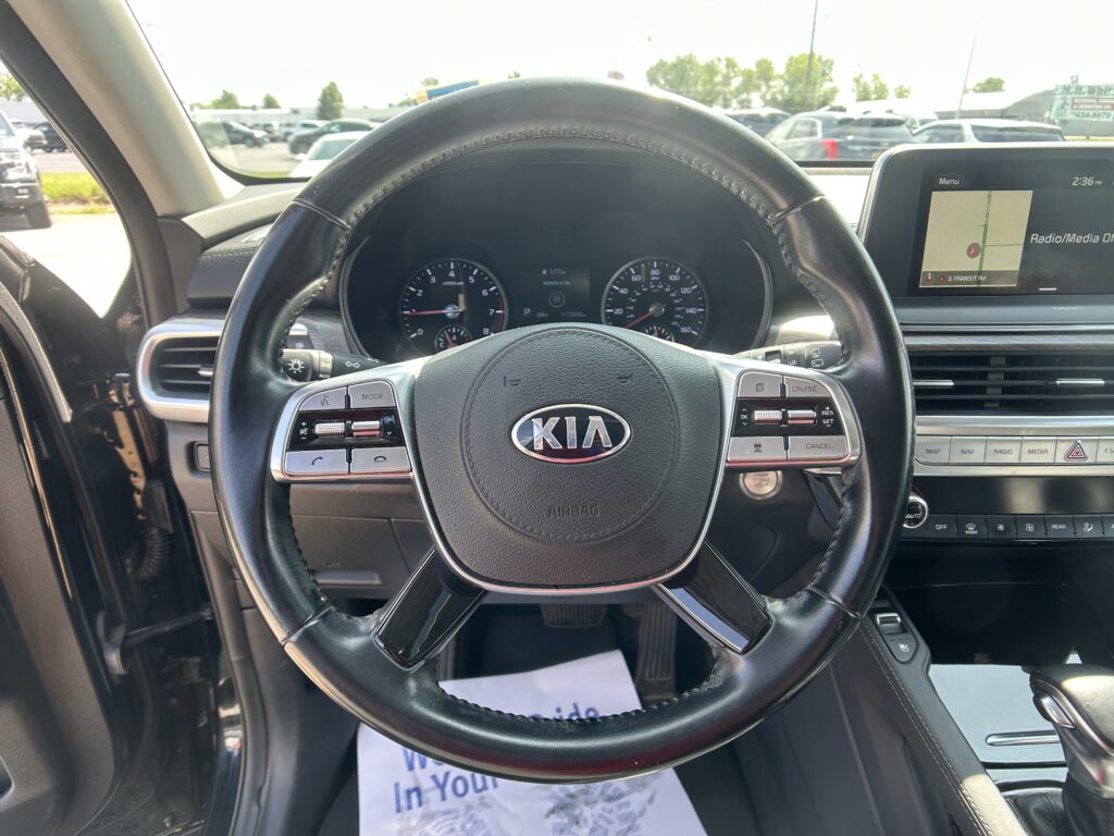 Driver's view of a Kia interior featuring a large steering wheel, instrument cluster, and a wide dashboard display screen in sunlight outside the windshield.