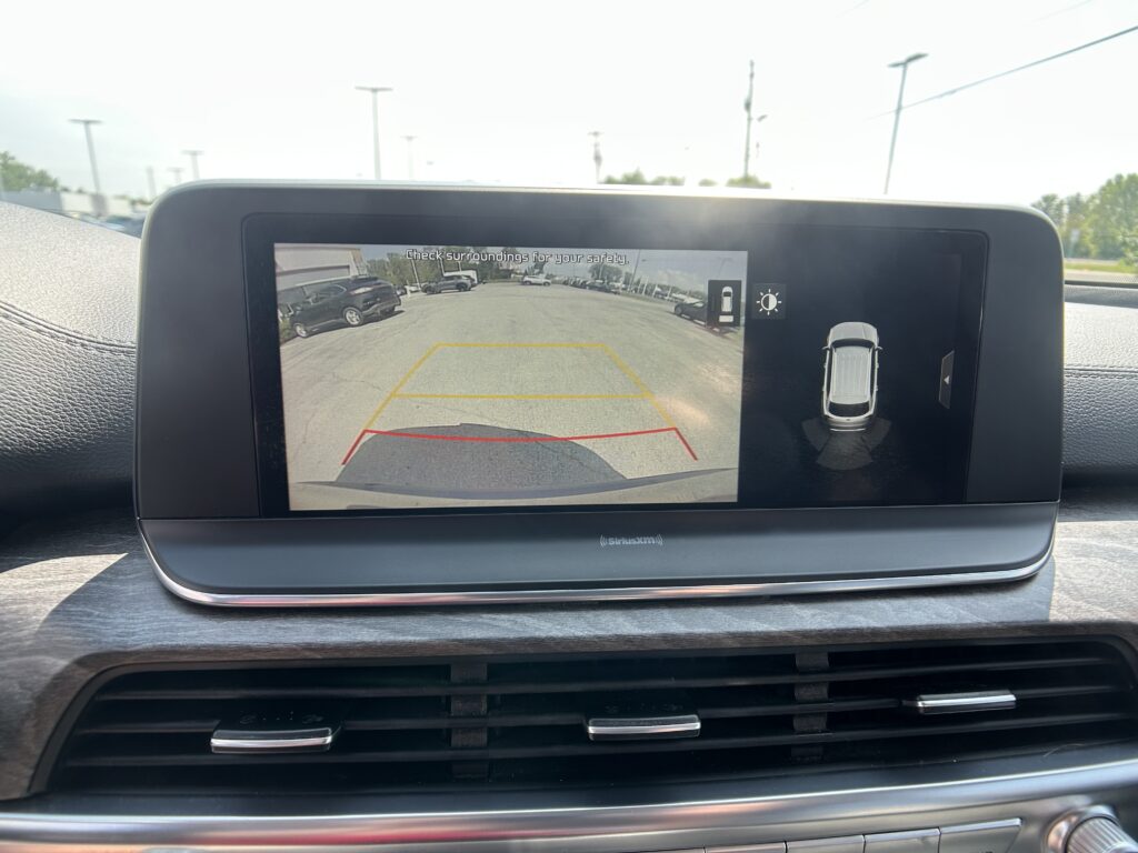 Rear-view camera display on a car dashboard showing a parking lot, with yellow and red guiding lines and the message 'Check surroundings for your safety.'